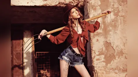 Fashionable woman in a brown blouse and denim shorts holding a pitchfork in a rustic, sunlit barn setting.