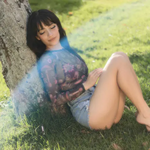 Young woman in a floral mesh top sitting by a tree in a sunny green meadow.