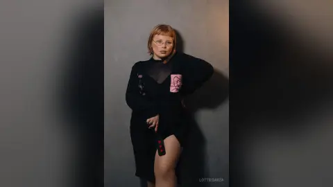 Redheaded woman in a black long-sleeve dress posing against a grey wall in a moody studio setting.