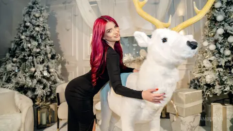 Woman in a black dress hugging a large white reindeer plush next to Christmas trees in a festive holiday setting.