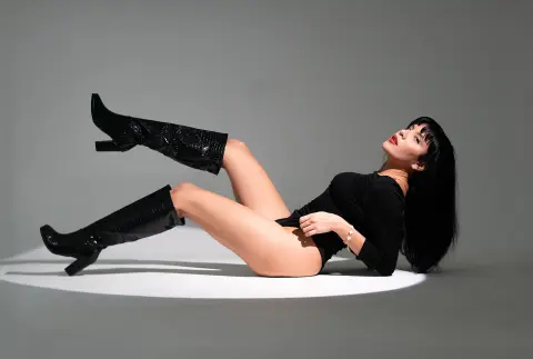 Stylish woman in a black bodysuit and tall patent leather boots posing on a studio floor with dramatic lighting.