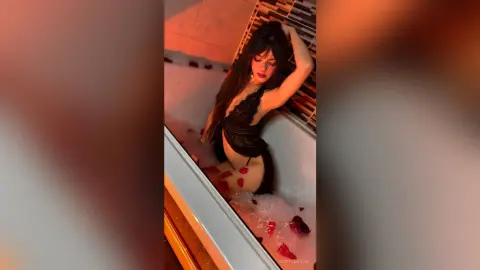 Glamorous woman in a black lace bodysuit relaxing in a bubble bath with rose petals in moody red lighting.