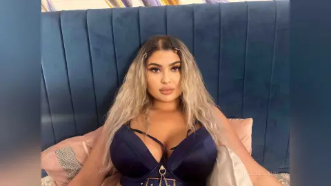 Brunette woman in a navy blue satin bodysuit relaxing against a blue velvet headboard in a cozy bedroom setting.