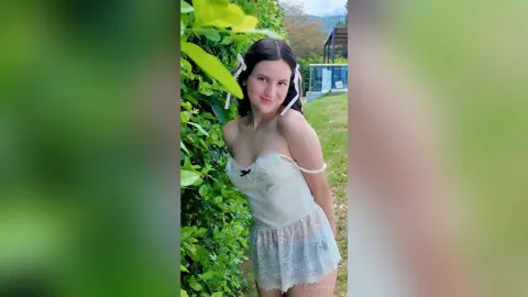 Smiling young woman in a white lace camisole outfit standing by lush green garden bushes outdoors.