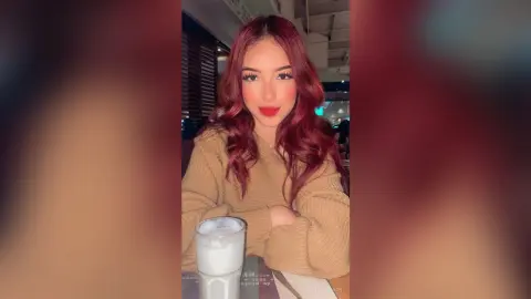 Young woman with wavy red hair wearing a tan sweater sitting at a restaurant table with a drink.