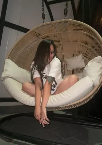 Young woman in a white t-shirt relaxing in a hanging wicker egg chair at night.
