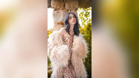 Elegant woman in a beige faux fur coat and leopard print dress posing outdoors in soft natural light.