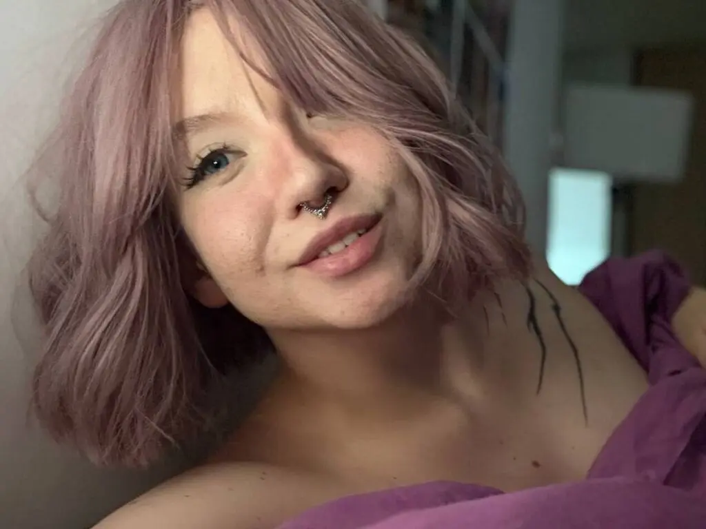 JoyceTreier smiles with her pink hair and nose ring, showing her neck and shoulder while wearing a purple top.