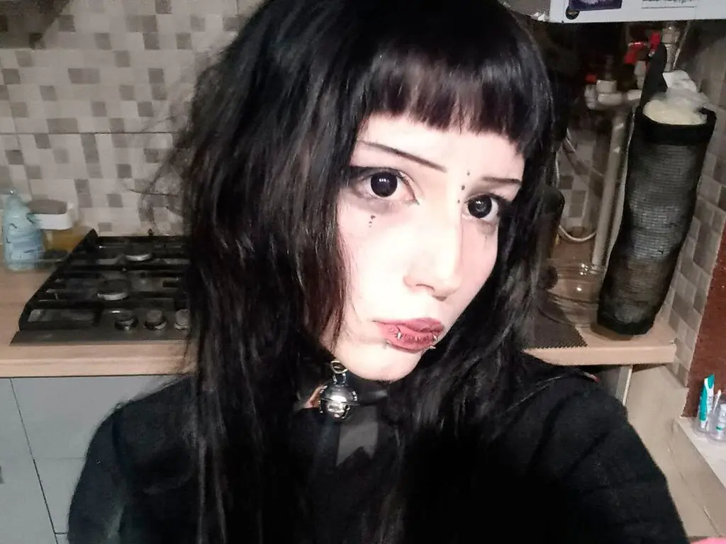 Goth woman with dark bangs and black outfit posing in a kitchen setting with a moody aesthetic.