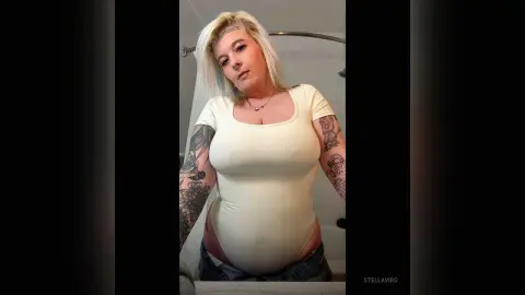Blonde woman in a white bodysuit posing in a bathroom with visible arm tattoos.