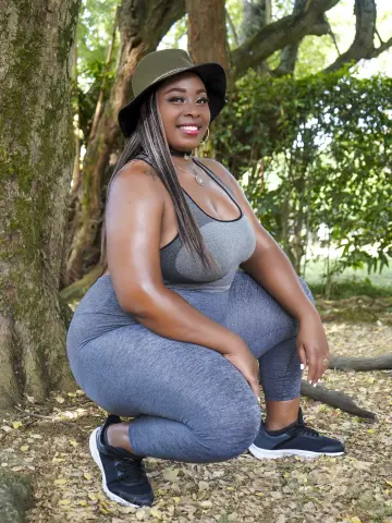 Smiling Black woman in grey yoga outfit and black sun hat posing in a lush green forest setting.
