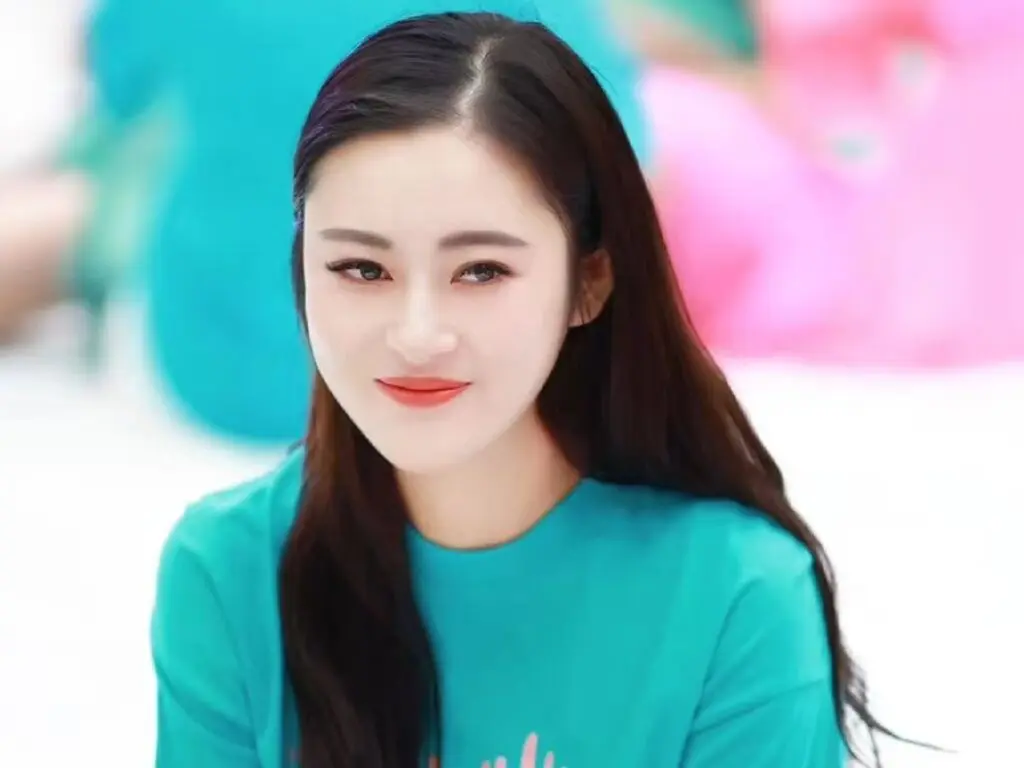 Smiling young woman with long dark hair wearing a teal t-shirt in a bright, soft-focus setting.