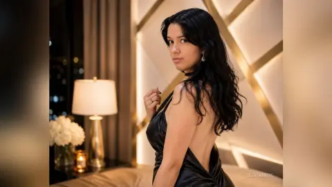 Elegant woman in a black satin backless dress posing in a luxury bedroom with warm ambient lighting.