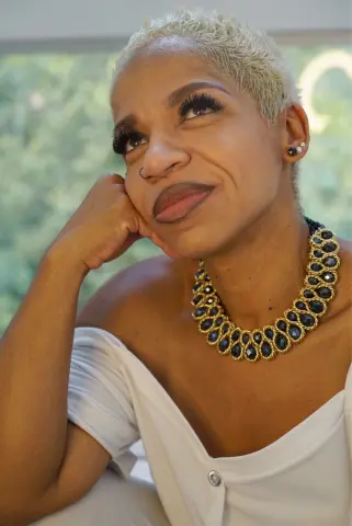 Smiling woman with short blonde hair wearing a white top and chunky gold statement necklace indoors.