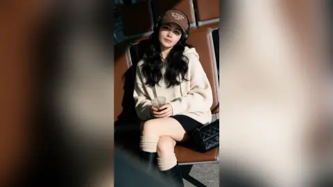 Young woman in a cozy cream sweater and black cap listening to music while sitting in an airport lounge.