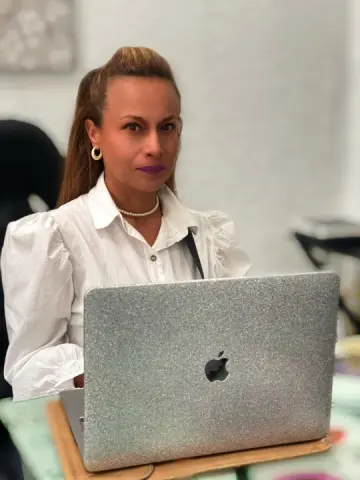 Professional woman in a white blouse working on a silver laptop in a bright office setting.