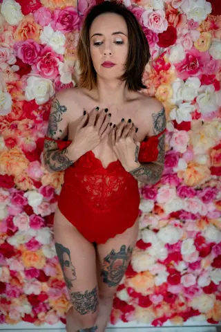 TaylaRae poses in a red lace bodysuit against a wall of pink and white roses, using her hands to touch her chest and cleavage.