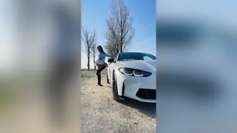 DyandraEve poses next to a white car on a dirt road, wearing a white skirt and black thigh-high boots.