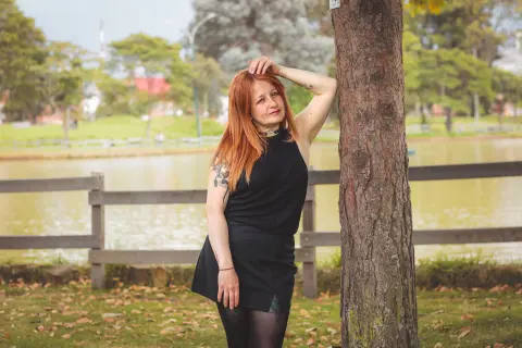 Redheaded woman in a black sleeveless dress leaning against a tree by a peaceful lake in a park.