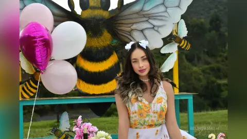 Smiling woman in a floral sundress posing in front of a giant bee sculpture and pink balloons outdoors.