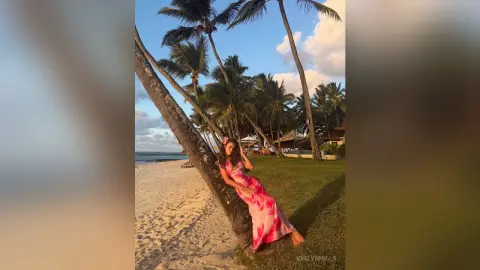 Woman in a pink floral maxi dress leaning against a palm tree on a tropical beach at sunset.