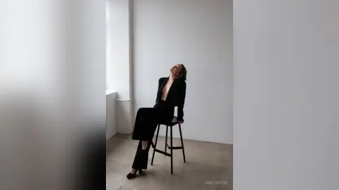 EmilyGiffin poses sitting on a stool in a black suit, leaning her head back against a white wall.