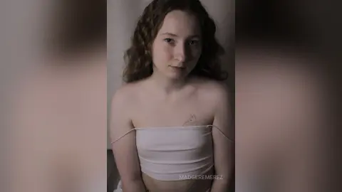 Young woman with wavy hair wearing a white tube top in a soft, moody indoor setting.