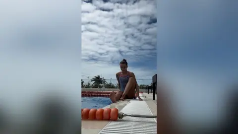Young woman in a pink swimsuit sitting by a swimming pool under a cloudy sky.