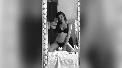 MontseFerrera takes a mirror selfie wearing black bra and panties while sitting on a white bed.