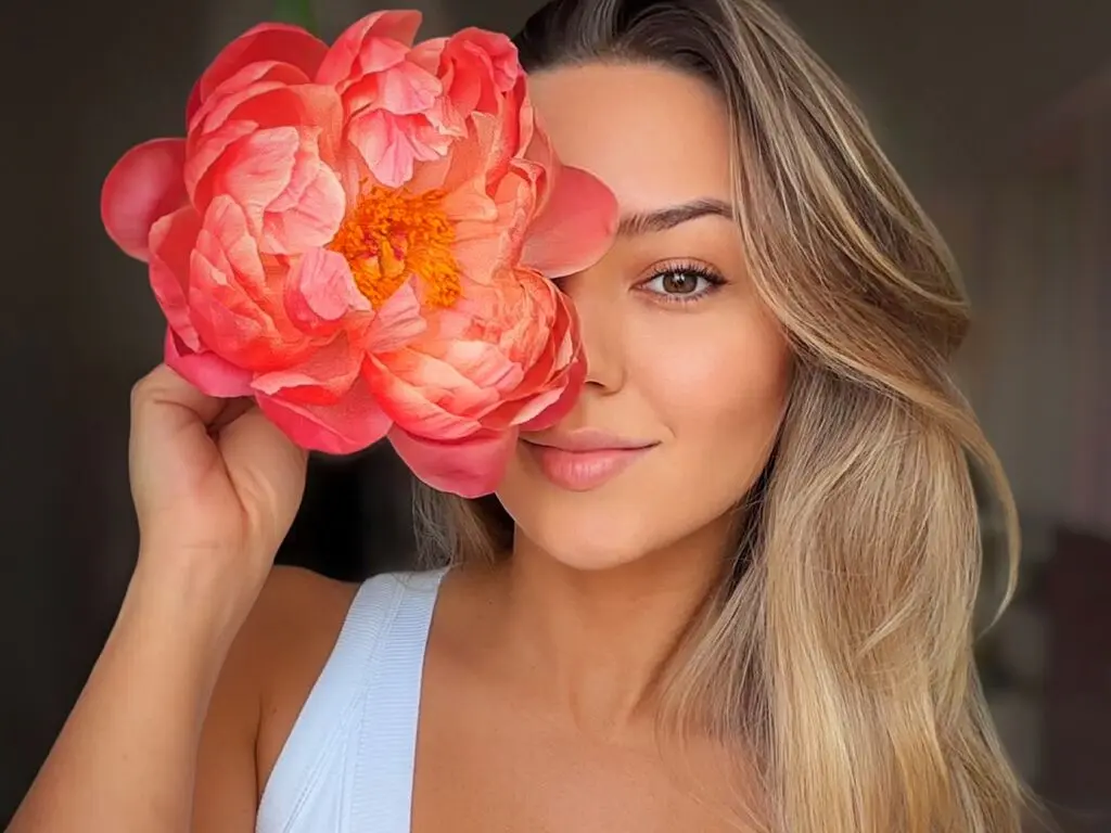 Smiling blonde woman in a white tank top holding a large pink peony over one eye for a dreamy aesthetic portrait.
