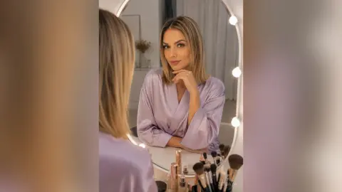 Blonde woman in a lavender silk robe looking into a lighted vanity mirror in a cozy, serene bedroom setting.