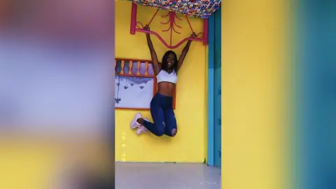 NatyGlow jumps in the air wearing a white crop top and blue jeans against a bright yellow wall.