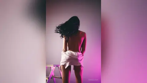 Back view of a woman with long dark hair in a white shirt against a moody pink and purple studio background.