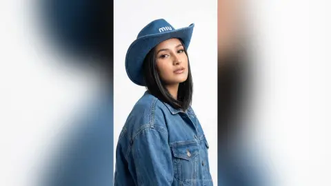 AlisonColemans poses in a blue denim jacket and a matching blue cowboy hat against a plain white background, looking toward the camera with dark hair.