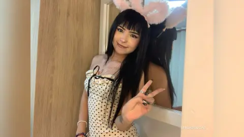 Smiling young woman with black bangs wearing a polka dot dress and pink bunny ears in a bright room.