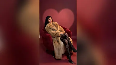 Elegant woman in a luxury faux fur coat and black lace tights sitting against a romantic red heart backdrop.