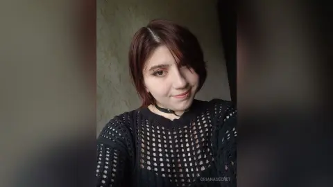 OrianaSecret — Young woman with short reddish hair wearing a black mesh sweater and choker in a moody indoor setting.