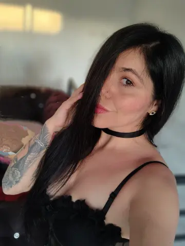 Smiling woman with long black hair wearing a black lace top and choker in a bright indoor setting.