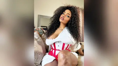 BossyBella — BossyBella poses in a white off-the-shoulder top and red corset, showing off her long, curly black hair and heavy eye makeup.