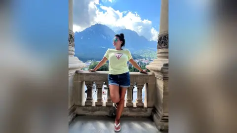 MissyJolie stands on a stone balcony wearing a light green t-shirt and denim shorts against a backdrop of mountains and clouds.