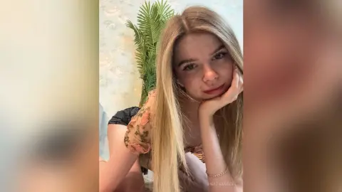 UnataMigdel is a blonde woman with long hair, posing with her chin resting on her hand while wearing a floral top.