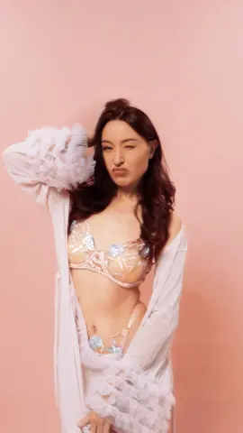 Playful woman in white ruffled robe and floral lace lingerie posing against a pink background.