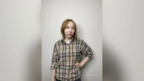 Young woman in a brown plaid button-down shirt posing against a plain white studio background.