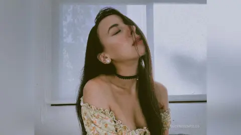 LucyRosenfelds poses with her eyes closed and head tilted back, wearing a floral off-the-shoulder top and a black choker.