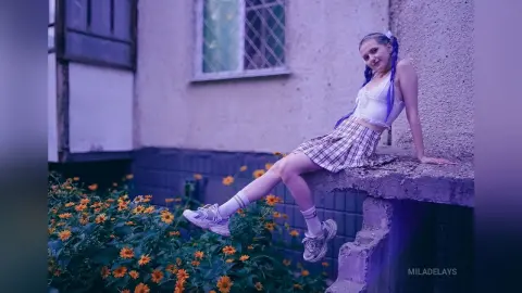 Young woman in a white crop top and plaid skirt sitting on a concrete ledge near garden flowers.