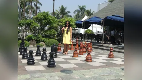 Woman in a yellow sundress standing by a giant outdoor chessboard in a sunny tropical garden setting.