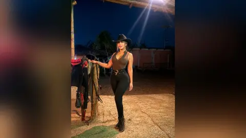 Stylish woman in black tank top, leggings, and cowboy hat posing outdoors at night near a rustic wooden post.
