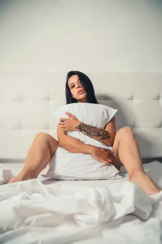 Tattooed woman in white bedding hugging a pillow in a bright, minimalist bedroom setting.