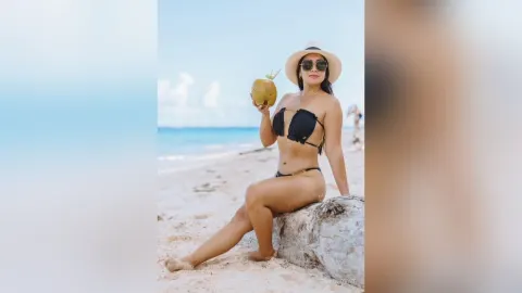 DemmiLovat — DemmiLovat sits on a log on a sunny beach wearing a black bikini and sunglasses while holding a coconut.