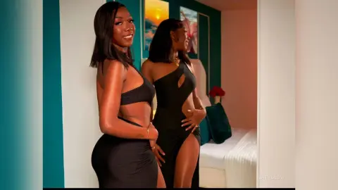 Smiling Black woman in a sleek black cutout maxi dress posing in a modern bedroom setting.